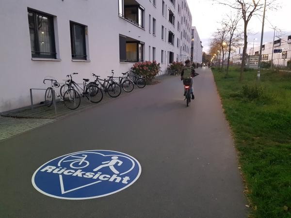 Auf einem asphaltierten Geh- und Radweg ist eine große blaue Bodenmarkierung mit den Symbolen für Fuß- und Radverkehr sowie dem Wort „Rücksicht“ zu sehen. Der Weg verläuft entlang eines Gebäudes und wird von einer radfahrenden Person genutzt. Die Markierung weist auf ein rücksichtsvolles Miteinander von Fuß- und Radverkehr hin.