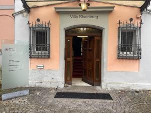 Das Foto zeigt den Eingang zu einem orangenen Haus. Mittig ist eine halbgeoöffnete, dunkelbraune Tür aus Holz, daneben sind links und rechts je ein Fenster.
