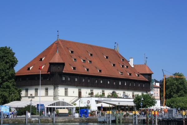 Auf dem Bild zu sehen ist das historische Konzilgebäude in Konstanz am Bodensee mit charakteristischem roten Ziegeldach und weißer Fassade. Das markante mittelalterliche Gebäude beherbergt heute eine Gaststätte und liegt direkt am Hafen mit Schiffsanlegestellen im Vordergrund. Aufnahme bei klarem Himmel an einem sonnigen Tag.