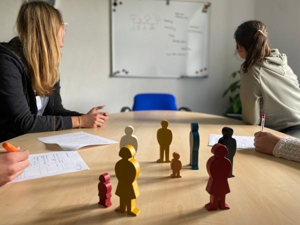 Auf dem Bild zu sehen ist eine Beratungssituation am Tisch mit zwei Personen, die Dokumente bearbeiten, während bunte Holzfiguren in der Mitte verschiedene Familienkonstellationen symbolisieren. Die Szene zeigt eine typische Beratungssituation im Bereich Familie und Soziales in einem hellen Büroraum. Im Hintergrund ist ein Whiteboard an der Wand zu sehen.