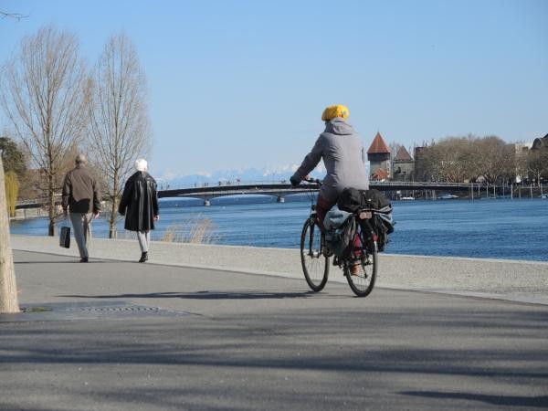 Radfahrerin mit gelber Mütze fährt auf der Seepromenade am Bodensee entlang, während zwei Fußgänger spazieren gehen. Im Hintergrund sind der blaue See, eine Brücke und historische Türme der Konstanzer Altstadt sowie die schneebedeckten Alpen am Horizont zu sehen. Aufnahme an einem klaren, sonnigen Tag.