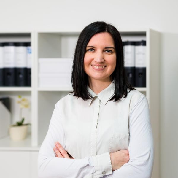 Frau mit schulterlangen dunklen Haaren; sie trägt eine helle Bluse und hat die Arme vor sich verschränkt; im Hintergrund ein Regal mit Aktenordnern