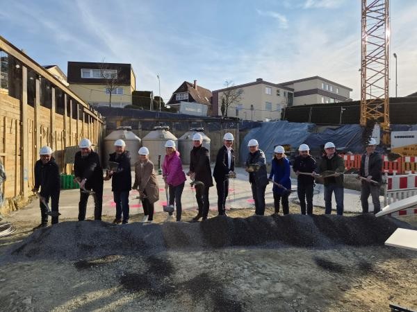 Spatenstich für die Grundschule Wollmatingen: Eine Gruppe von zwölf Personen mit weißen Bauhelmen steht mit Spaten auf einer Baustelle und symbolisiert den Baubeginn. Im Hintergrund sind Wohngebäude, ein Baukran, Baucontainer und eine Holzverschalung zu sehen.