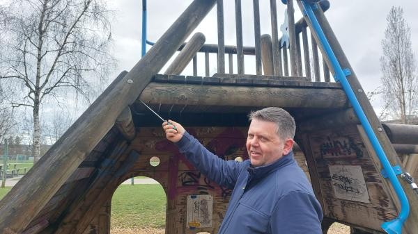 Ein Mann in einer blauen Fleecejacke steht vor einem großen Holzspielgerät in Form eines Schiffs auf einem Spielplatz. Er hält einen Schraubendreher in der rechten Hand und sticht damit in eine Holzverbindung an der Dachkante des Geräts, um das Holz auf Fäulnis zu prüfen. Im Hintergrund sind kahle Bäume, eine Grünfläche und ein bewölkter Himmel zu sehen. Das Holzgerät weist Graffiti auf.