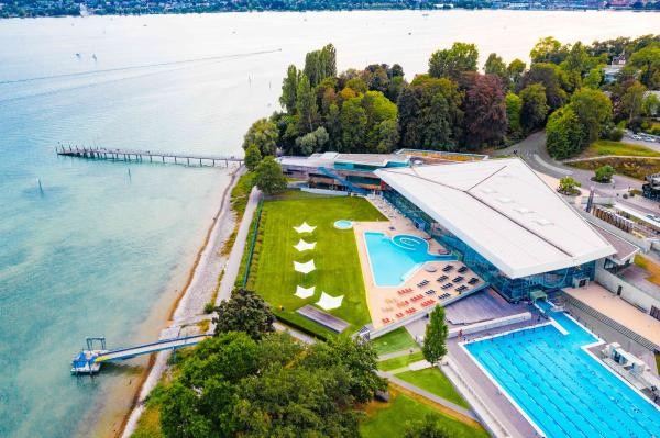 Ein Gebäude mit grüner Wiese, zwei Schwimmbecken und dem Bodensee von oben