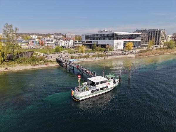Luftaufnahme: Ein weiß-grünes Wasserbusschiff liegt an einem Steg am Konstanzer Bodenseeforum. Zahlreiche Menschen warten auf dem Steg. Im Hintergrund sind das Bodenseeforum, weitere Gebäude und der belebte Uferpark bei strahlendem Frühlingswetter zu sehen.