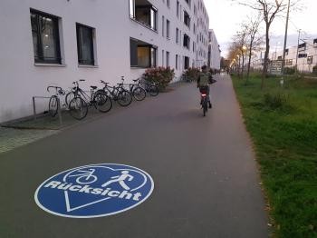 Zu sehen ist ein asphaltierter Geh- und Radweg entlang einer Bahnlinie. Auf dem Boden befindet sich eine blaue Markierung mit den Symbolen für Fuß- und Radverkehr sowie dem Hinweis „Rücksicht“. Im Hintergrund fährt eine Person mit dem Fahrrad, daneben stehen abgestellte Fahrräder vor einem Gebäude.