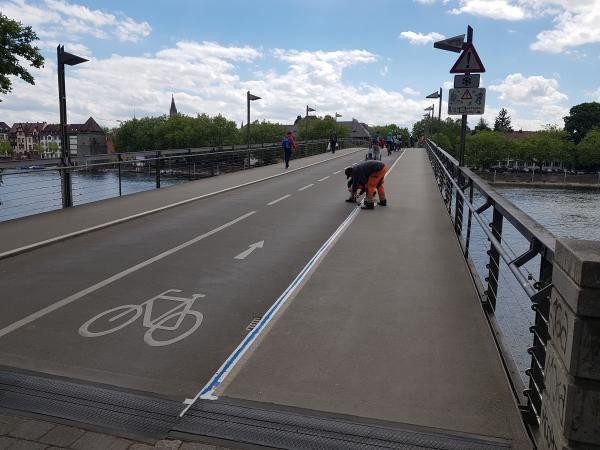 Radweg auf einer Brücke mit blauer Markierung und Fahrradsymbolen am Boden. Mitarbeitende in orangefarbenen Warnwesten arbeiten bei einem Schild mit Gefahrenhinweisen, im Hintergrund radeln mehrere Personen auf dem Weg. Rechts ist das Ufer mit Wasser zu sehen, im Hintergrund Gebäude und Kirchturm.