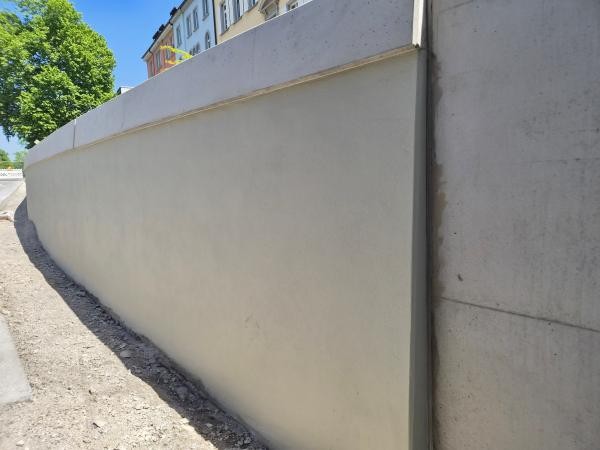 Helle, frisch verputzte Betonwand an einer Straße, aufgenommen aus schräger Perspektive. Oben sind Teile angrenzender Wohnhäuser und ein grüner Baum zu sehen.