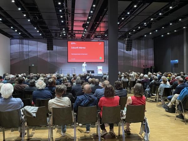 Blick von hinten auf ein sitzendes Publikum in einem Saal; im Hintergrund ein Sprecher auf einer Bühne