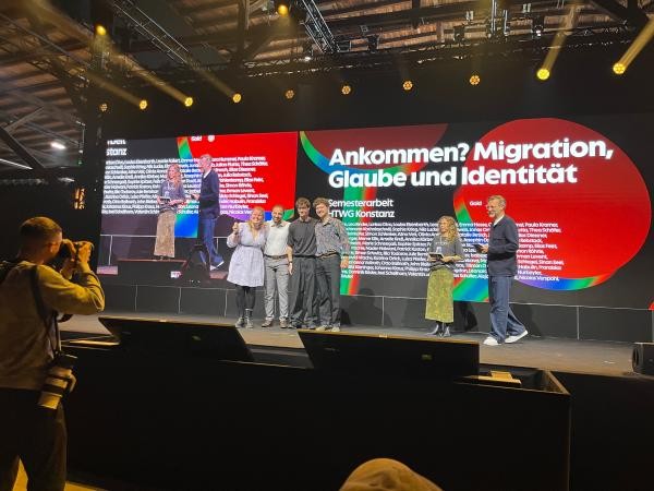 Eine Personengruppe steht auf einer Bühne. Im Hintergrund ist projizierter Text zu sehen: "Ankommen? Migration, Glaube und Identität"