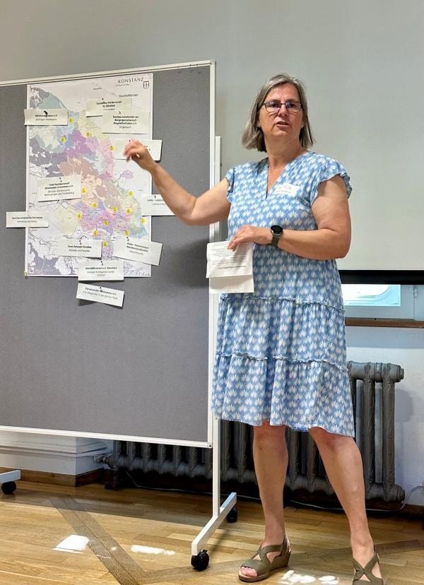 Das Foto zeigt eine Frau mit blonden, kinnlangen Haaren, Brille und hellblauem Kleid. Sie steht vor einer Flipchart-Wand mit weißem Papier und und bunten Karten.