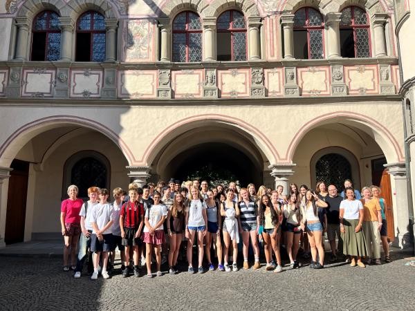 Copyright: Stadt Konstanz Auf dem Foto ist eine große Gruppe Jugendlicher in sommerlicher Kleidung in einem Innenhof zu sehen. Im Hintergrund steht ein großes Gebäude aus Stein mit Bögen. Die Sonne scheint auf die Gruppe.