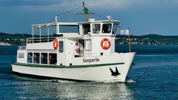 Das Fahrgastschiff „Seeperle" mit weißem Rumpf und Sonnendeck fährt auf dem Bodensee. Ein Besatzungsmitglied steht am Bug, im Hintergrund sind das Ufer und Hügel zu erkennen.