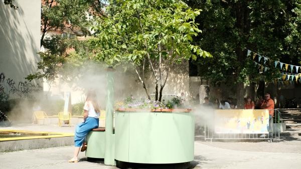 Der neue Sommerort Augustiner an einem sonnigen Tag: Eine Person sitzt auf einer runden hellgrünen Sitzbank unter einem jungen Baum in einem Pflanzkübel, aus dem eine kühlende Wassernebeldüse sprüht. Im Hintergrund stehen Menschen an einer Absperrung mit Informationsbanner, geschmückt mit bunten Wimpelketten. Foto: Raphael Rössler.