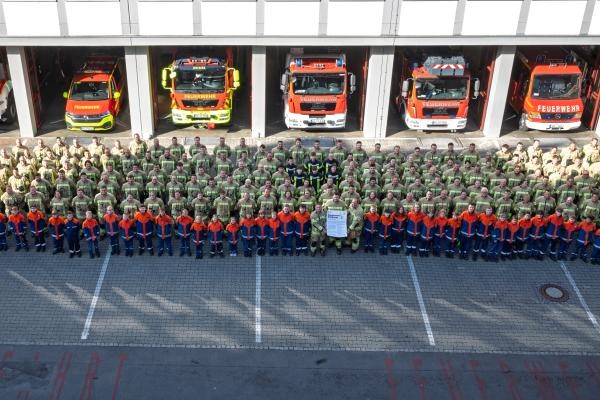 Feuerwehr Konstanz mit Konstanzer Erklärung Das Foto zeigt eine große Menschenmenge in dunkelgrüner Uniform mit gelben Warnstreifen und Helmen. Die Menge steht vor einem langen, flachen Garagen-Gebäude und guckt leicht schräg nach oben.