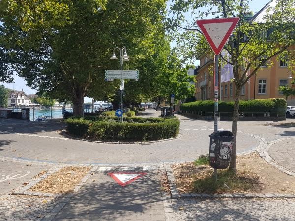 Neu markierte Kreuzung an der Fahrradbrücke in Konstanz: Am Boden ist ein rot-weißes Vorfahrt-gewähren-Dreieck aufgemalt, ergänzt durch ein entsprechendes Verkehrsschild am Straßenrand. Im Hintergrund sind Wegweiser zu Radolfzell, Kreuzlingen und der Altstadt sowie das Ufer des Bodensees mit Bäumen zu sehen.
