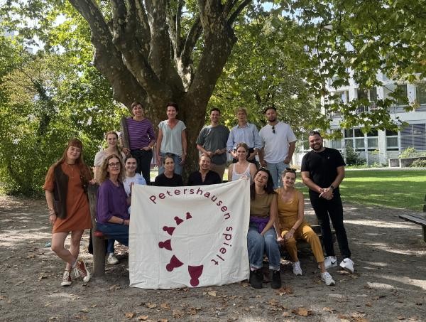 Petershausen spielt 15 Personen sitzen oder stehen verteilt unter einem Baum. Zwei von ihnen halten ein großeses Stoffbanner mit der Aufschrift "Petershausen spielt" darauf zentral nach oben.