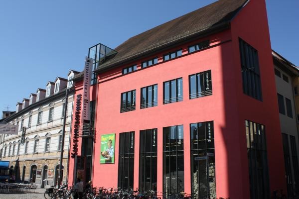 Das Kulturzentrum am Münster mit auffälliger pinkroter Fassade und großen Schriftzügen „Stadtbücherei" an den Fensterfronten. Vor dem Gebäude stehen zahlreiche Fahrräder, Passanten gehen über den Platz an der Wessenbergstraße.