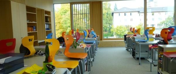 Leeres Klassenzimmer einer Konstanzer Schule mit bunten Stühlen in Orange, Gelb, Rot und Blau, die hochgestellt auf den Tischen stehen. Große Fensterfronten geben den Blick auf herbstliche Bäume und umliegende Gebäude frei, links befinden sich Holzregale mit Ordnern.
