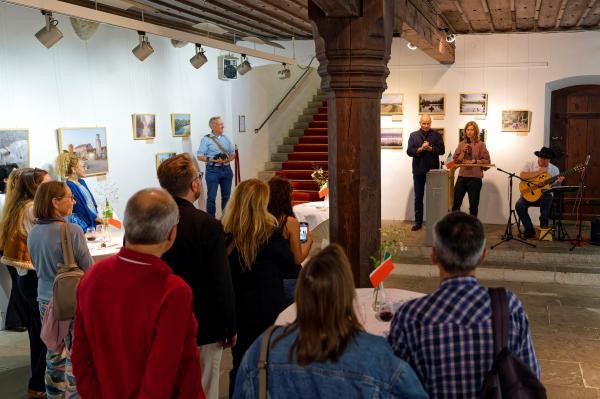 Begegnungswochenende: Lodi zu Gast in Konstanz Auf dem Bild sind einige Personen in einem historischen Raum, während einer Ausstellungseröffnung zu sehen. Sie blicken zu zwei Personen, die eine Rede halten. Rechts ist ein Musiker mit Gitarre zu sehen. An der Wand hängen Bilder.