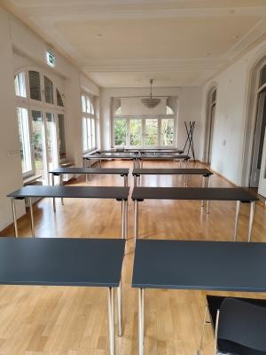 Das Foto zeigt einen Hauseingang mit geöffneter, dunkler Holztür. Die Hauswand ist orange. Neben der Tür befinden sich zwei vergitterte Fenster, über der Tür steht der Schriftzug Villa Rheinburg.