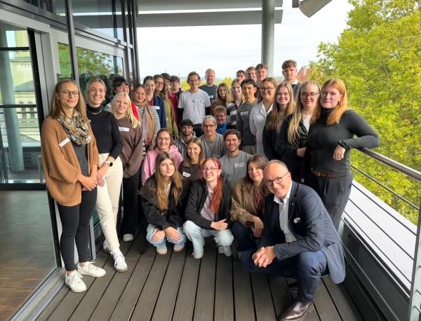 Eine Gruppe von Menschen steht (teilweise in der Hocke) auf einer Terasse im Hintergrund ist ein Baum