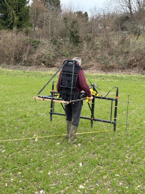 Mann mit einem am Rücken festgeschallten geomagnetischen Vermessungsgerät auf einer Wiese