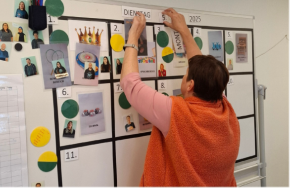 Eine Frau in orangefarbener Weste steht vor einem großen, beschrifteten Wochenplan an der Wand und bringt neue Aufgabenblätter an.