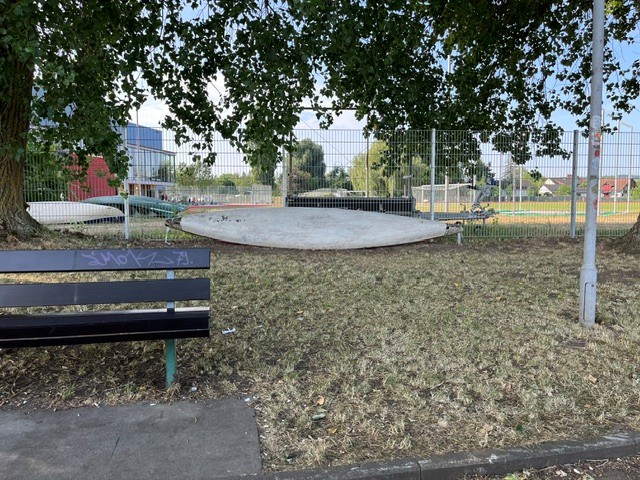 Mehrere SUP-Boards liegen unerlaubt an einem Metallzaun auf einer Grünfläche, im Vordergrund steht eine schwarze Parkbank mit Graffiti.