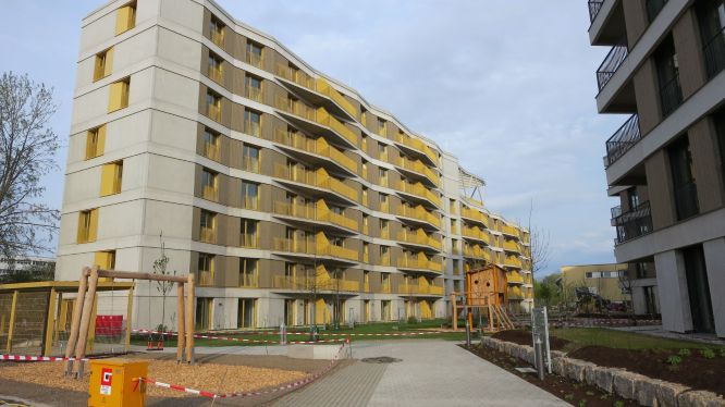 Modernes mehrgeschossiges Wohngebäude im Quartier Weiherhof mit gelben Balkonelementen und geschwungener Fassade. Im Vordergrund ein neu angelegter Innenhof mit Spielgeräten und Grünflächen.